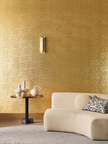 Casamance behang Métal pyrite patroon in goud met een zwarte tafel gouden lampen en een creme bank met daarop wit zwarte kussens